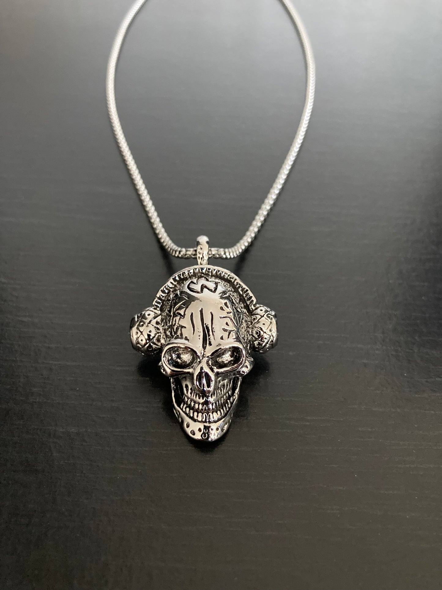 A close up view of a skull head wearing headphones pendant. This has a very detailed face including sunken eyes and an evil wide grin baring teeth. A silver colour chain holds the item which looks and feels good quality.