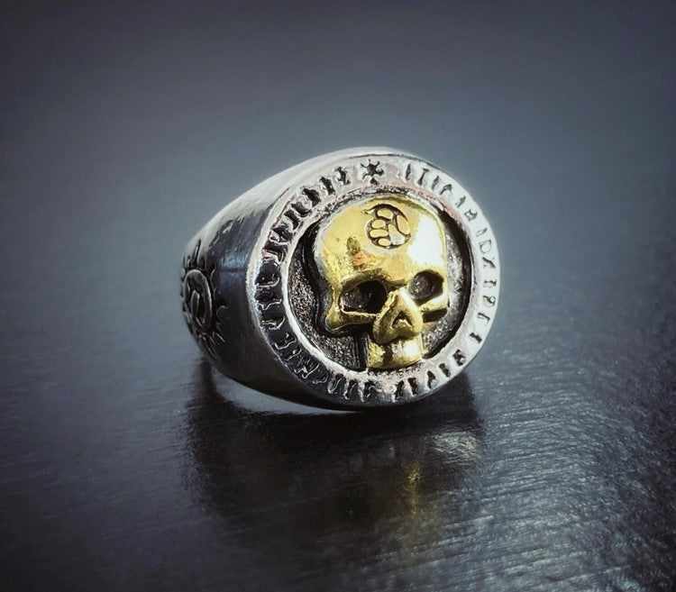 On a black surface is a silver coloured ring which is chunky looking. A gold coloured skull is imprinted on the main part of the ring. There is an engraved symbol just seen on the side of it.