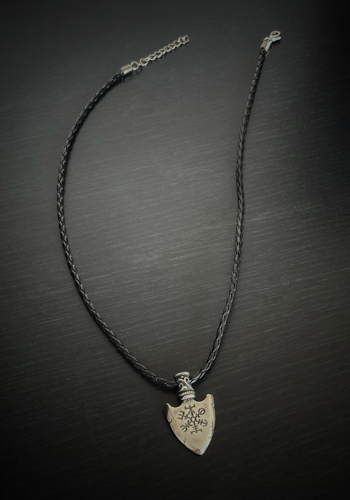 A full view of a black corded necklace with clasp for fastening at different lengths. The pendant to silver coloured in the shape of an arrow with a vegvisir design engraved onto it. 