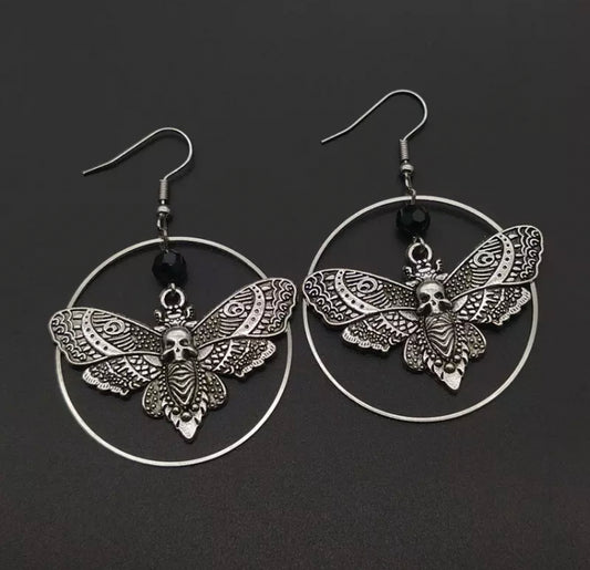 A pair of earrings that are thin hoops with death moths hanging in the centre of them. There is a hook to go into the earlobe and they are silver in colour. The moth itself has a stunning design etched onto the wings and a skull face in the middle part.