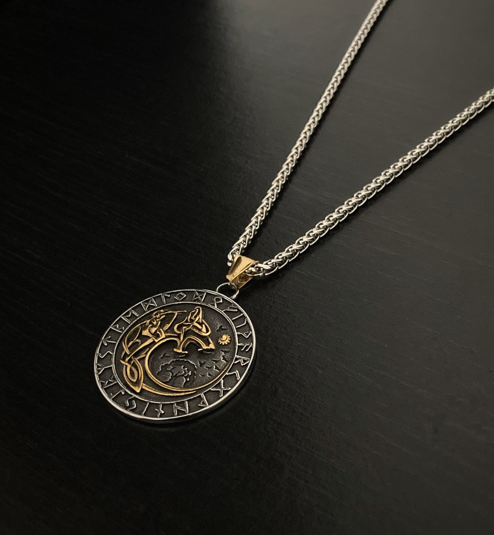 On a plain black surface is a stainless steel pendant on a chain of the same material. Lying at a slight angle you can see the details of the wolf inscription which features in the middle of the pendant whilst there are rune symbols adorning the edge