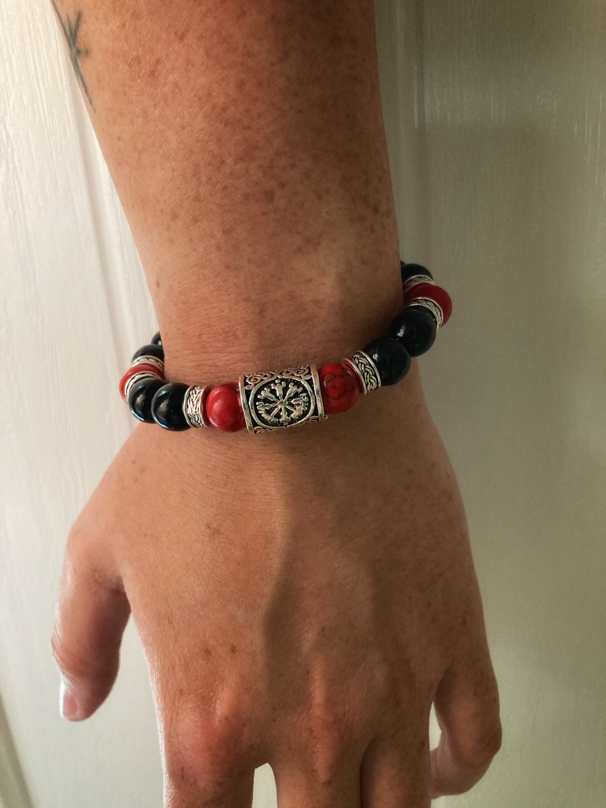 A hand is dangling down with a bead bracelet on the wrist. This features a helm of awe bead at the front with red turquoise beads as well as black ones making up the rest of the bracelet with a few metal patterned pieces throughout.