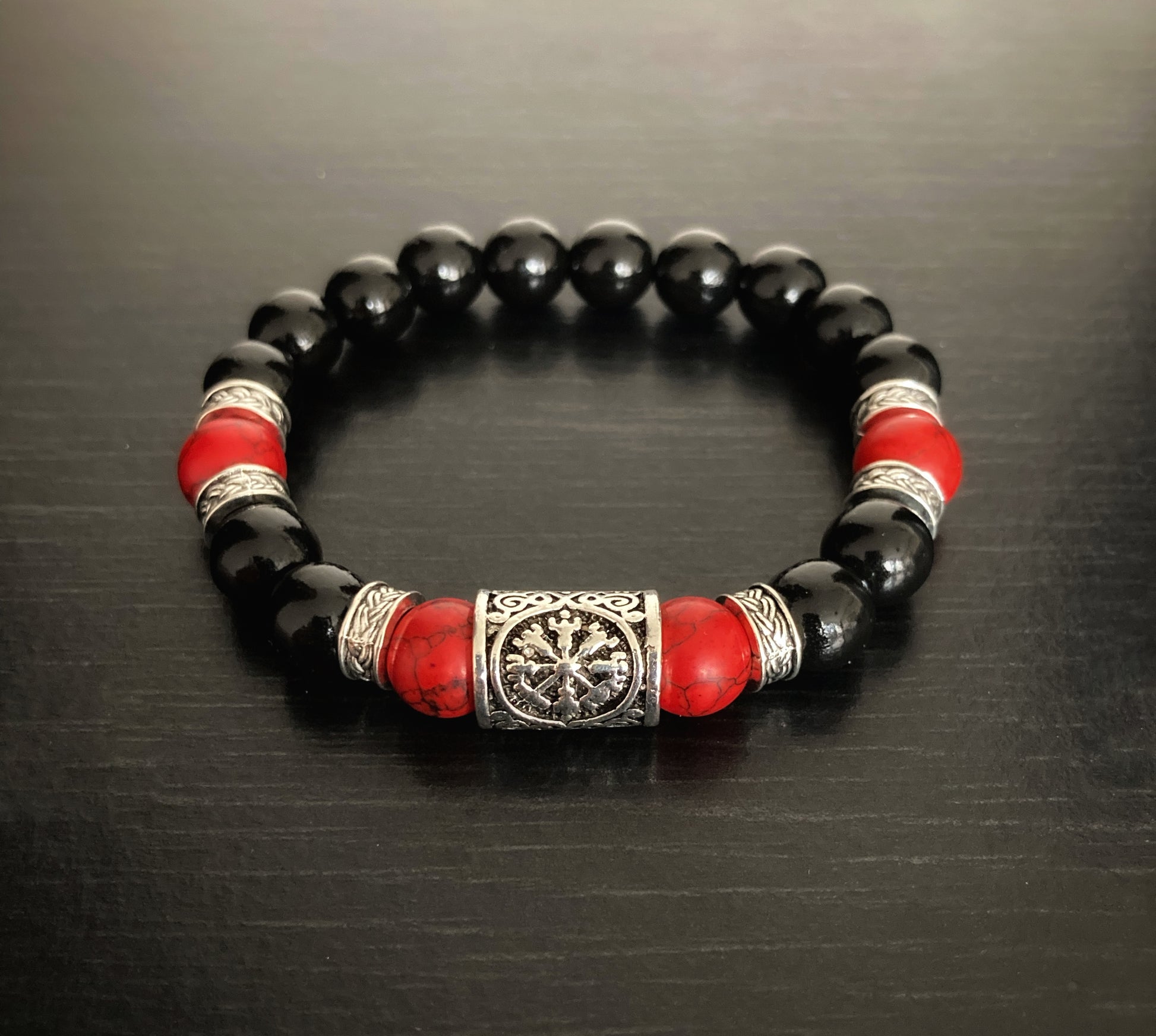 A stunning bracelet sits on a black background. This item has black wooden beads as well as red turquoise ones and between them are round silver metal patterned pieces. The main feature of it is a rune helm of awe also in silver.