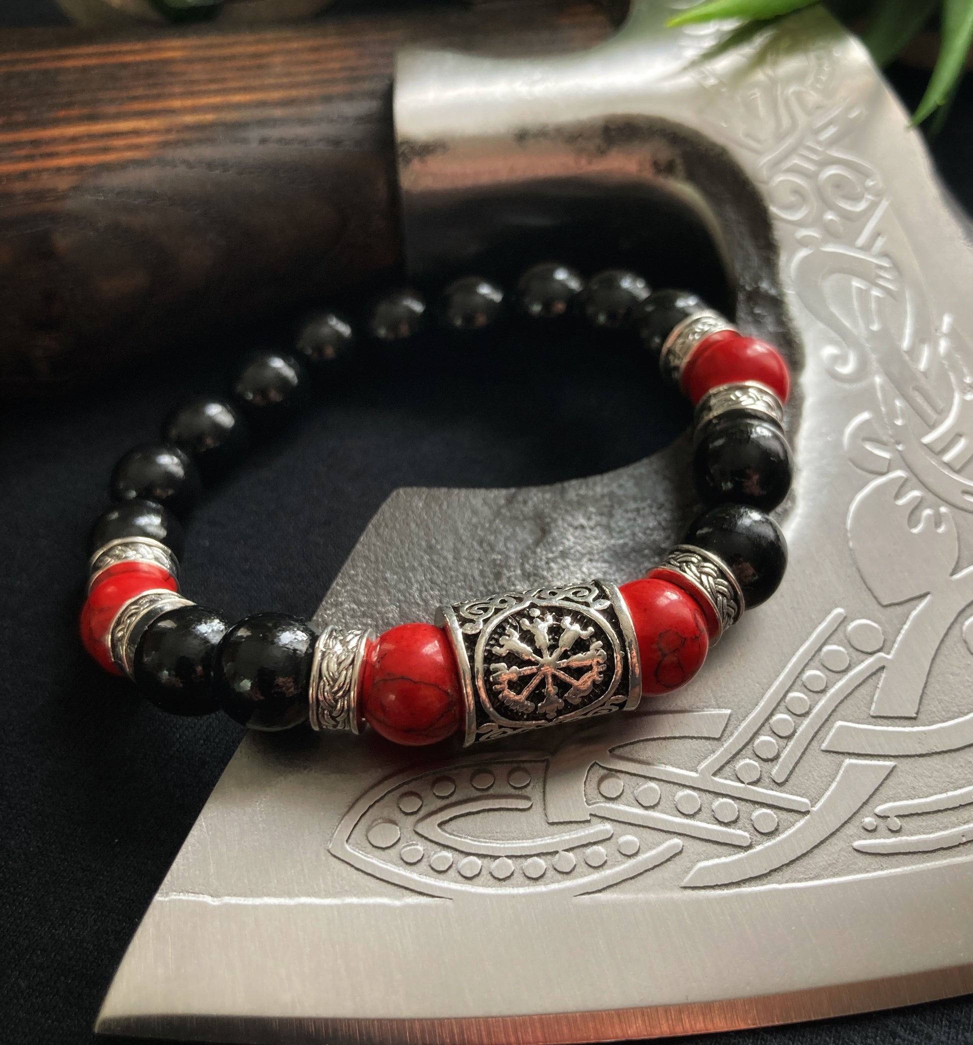 Draped over an axe blade lies a beautiful beaded bracelet. There are black wooden beads and red with turquoise running through them as well as narrow silver coloured patterned metal pieces. The main fetaure is a metal rune with an engraving.