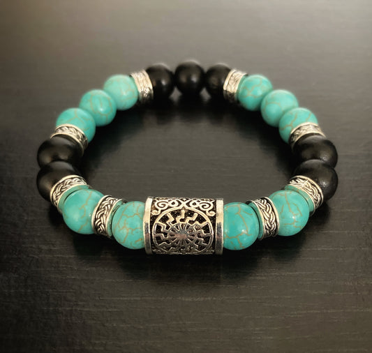 A bracelet made up of beads sits on a dark surface. There are turquoise and black beads going round the bracelet with small metal ring pieces between some of them. The main feature is a silver coloured metal rune with markings engraved on it.