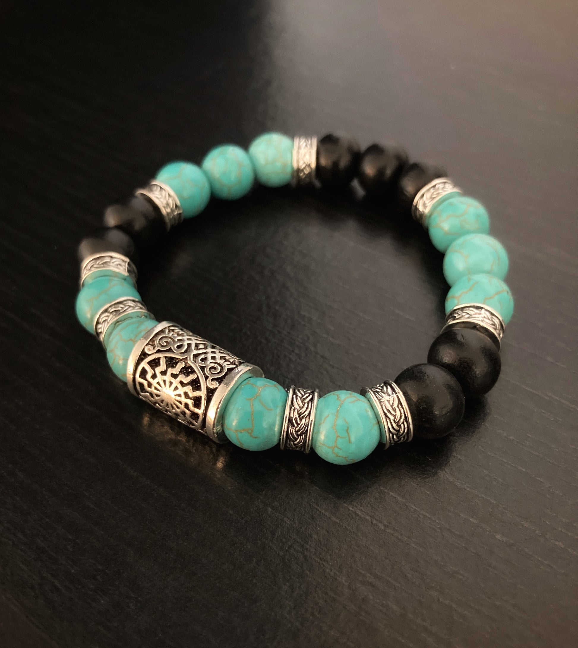 A turquiose and black beaded bracelet with a metal rune sits on a black surface. There are 7 of the black beads and 10 of the turquoise.