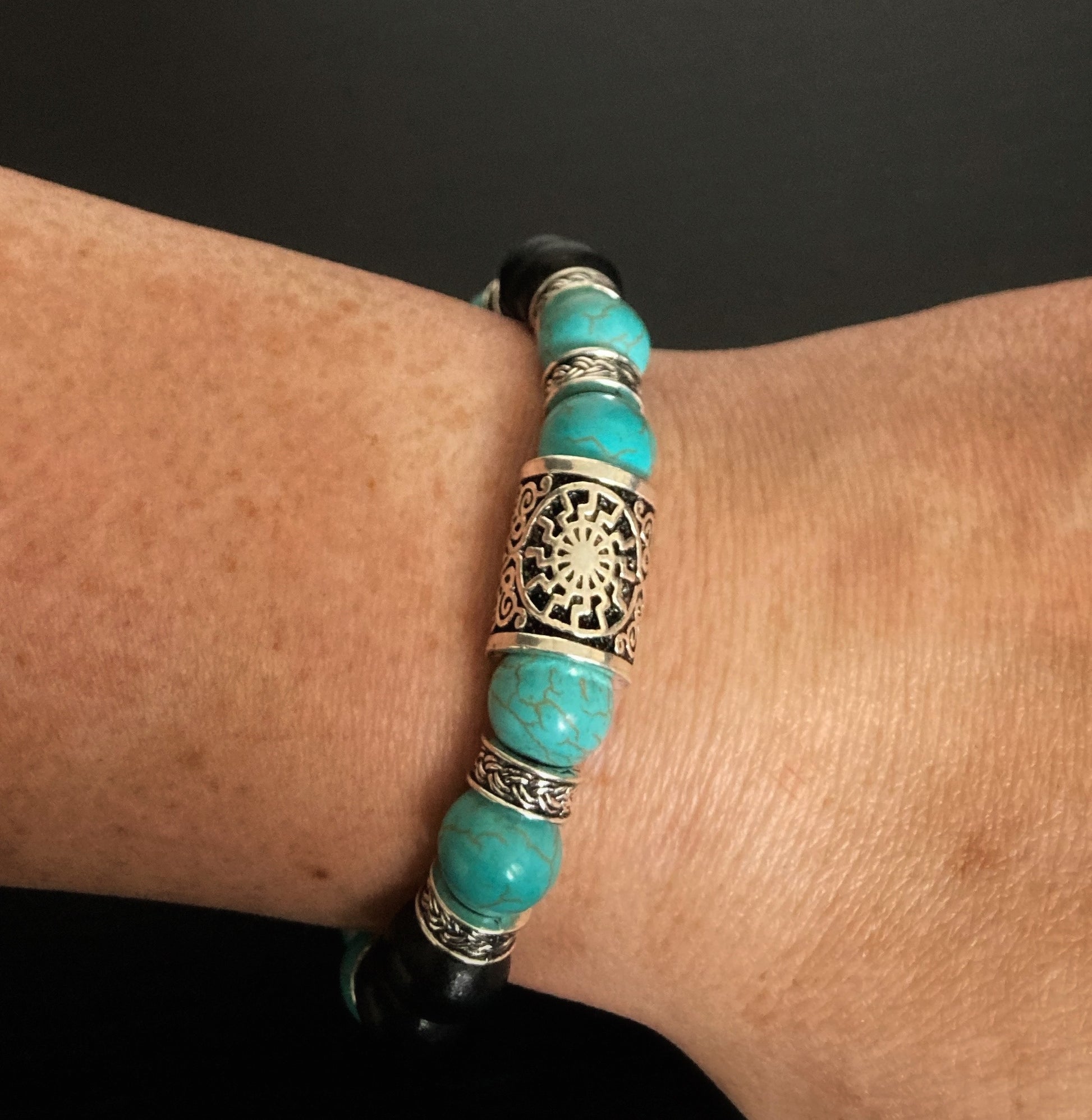 Sitting on a wrist is a beaded bracelet. You can see the main part which is a silver coloured metal rune with markings. Either side of that are turquiose wooden beads with a crackling pattern running through them. Between these beads are small metal ring shapes with patterning.