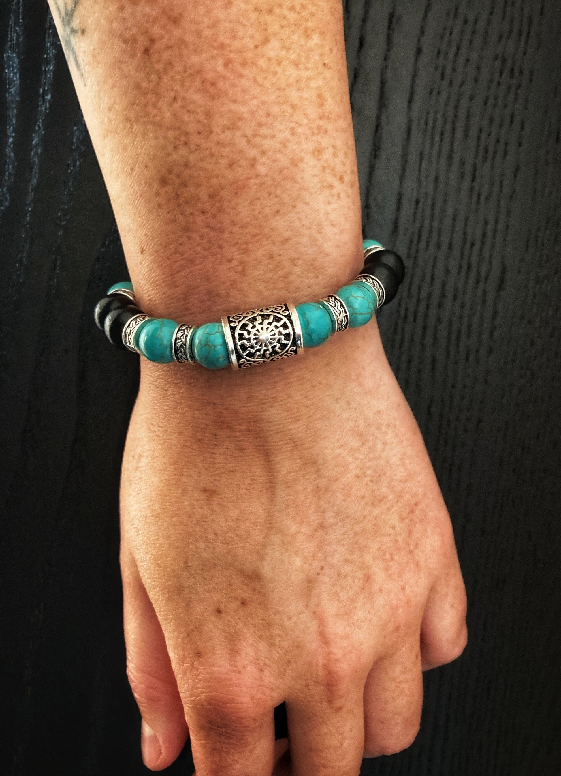 A arm dangles showing off a bracelet on the wrist. You can see the shiny silver coloured rune with a black sun symbol engraved onto it and the patterned thin metal ring pieces that seperate the beads.