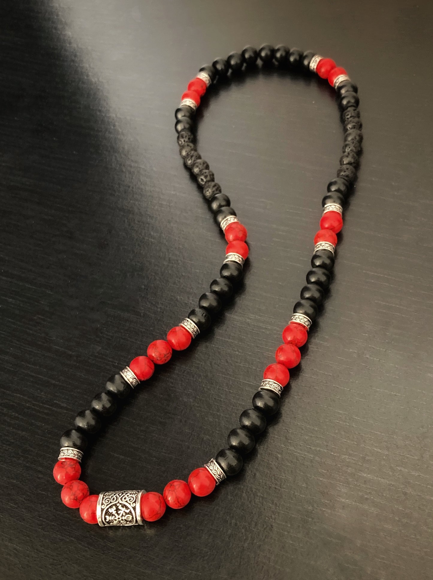 On a black surface sits a necklace made of beads. There are both black and red beads and these are broken up around the item with small metal ring shapes that are patterned.