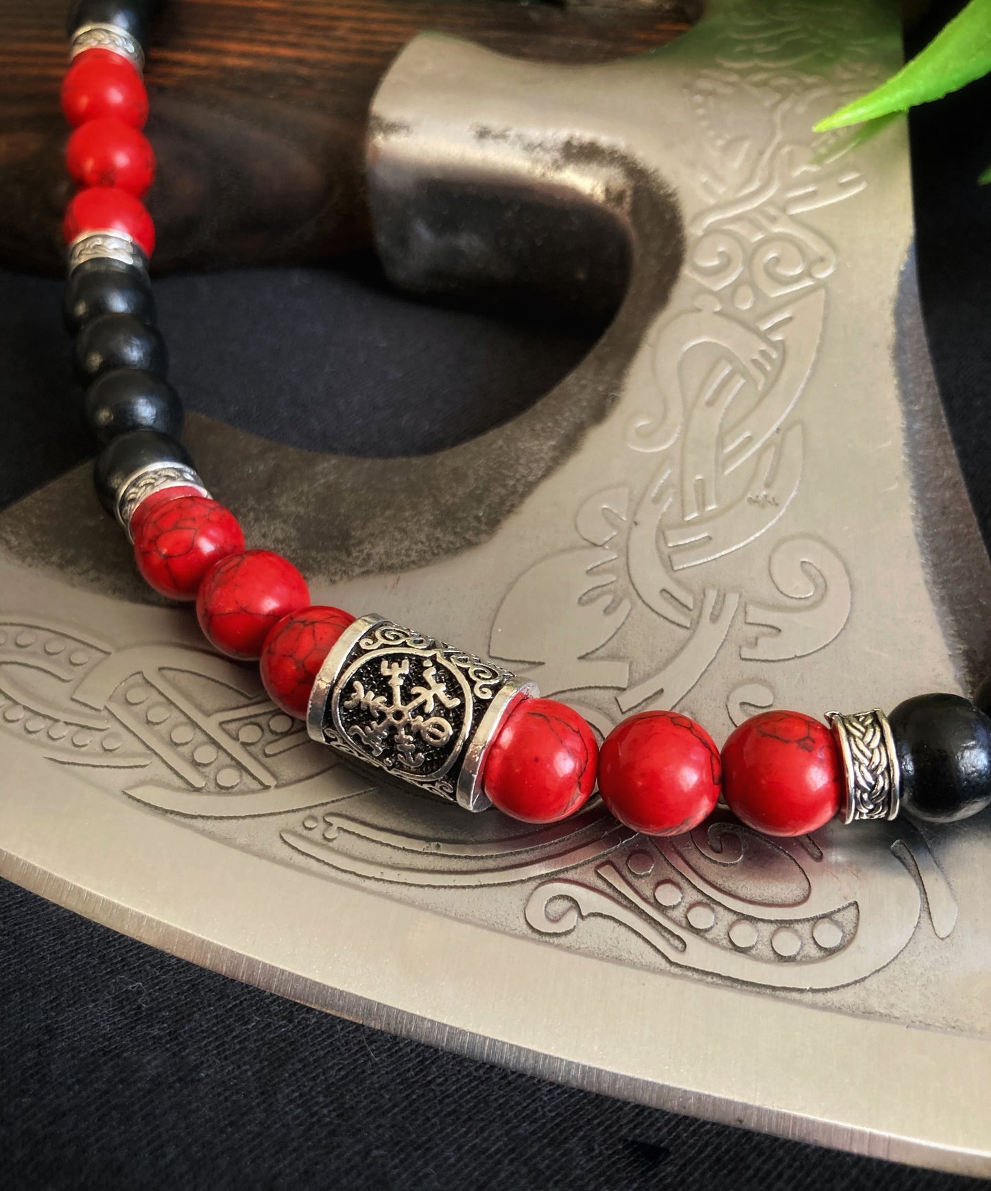 Part of a necklace sits on a axe blade. The vegvisir cylindrical metal bead is seen and is shiny in appearance. Both the red and black beads are glistening where the light is bouncing off of them.