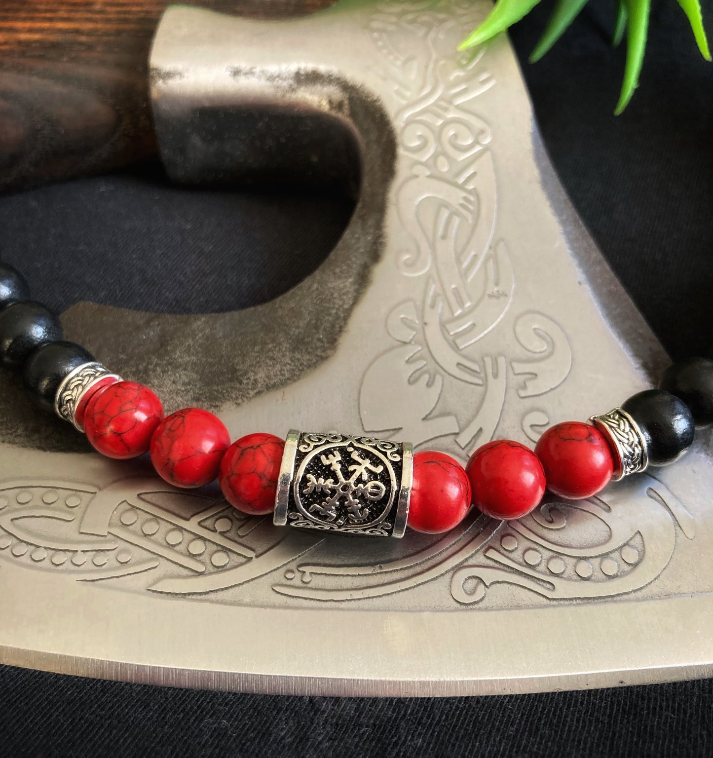Upon a axe blade sits a small section of a beaded necklace. You can see in detail the metal rune with its viking compass design on it. Either side of this are red shiny beads that have a darker colour running through them.