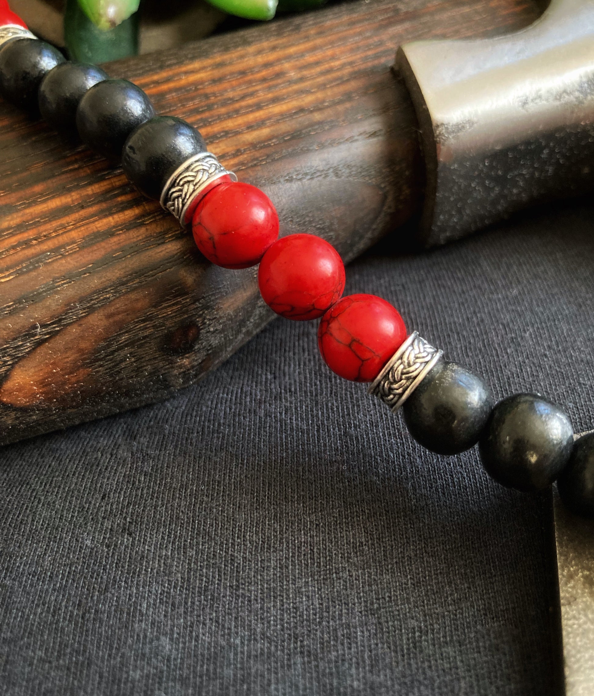 A section of a necklace is on show draped over an axe handle. You can see the red coloured beads that have a darker streak going through them. There are 3 of these beads and either side sits narrow metal bands to seperate the next part of which is black wooden style beads.