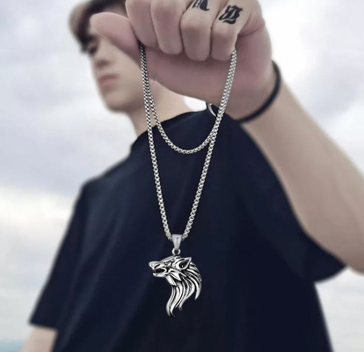 A teenage boy is holding a necklace in his left hand. The sturdy looking chain is in his clenched fist with a wolfs head pendant dangling from it. This is shiny in appearance and looks solid with detailed facial expressions to make it look ferocious.