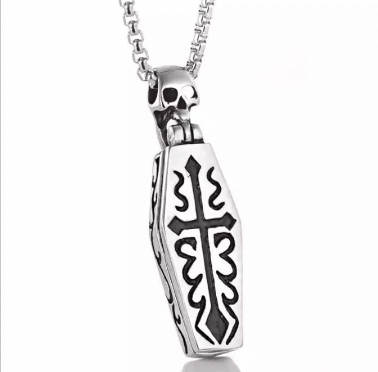 A pendant in the shape of a coffin is attached to a chain. Both are silver in colour and the coffin has a black cross engraved onto it and a skull at the top.