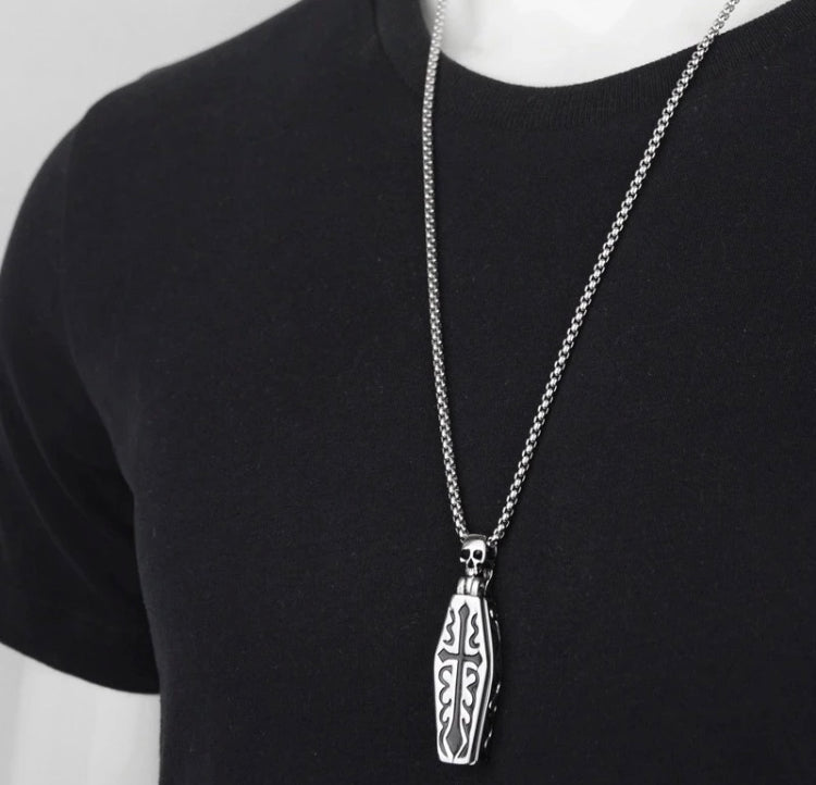 Around a neck against a black t-shirt sits a coffin pendant. This faces downwards with a cross engraved onto the outside of it in black and a skull features at the top of it with a chain feeding into either side of the head.