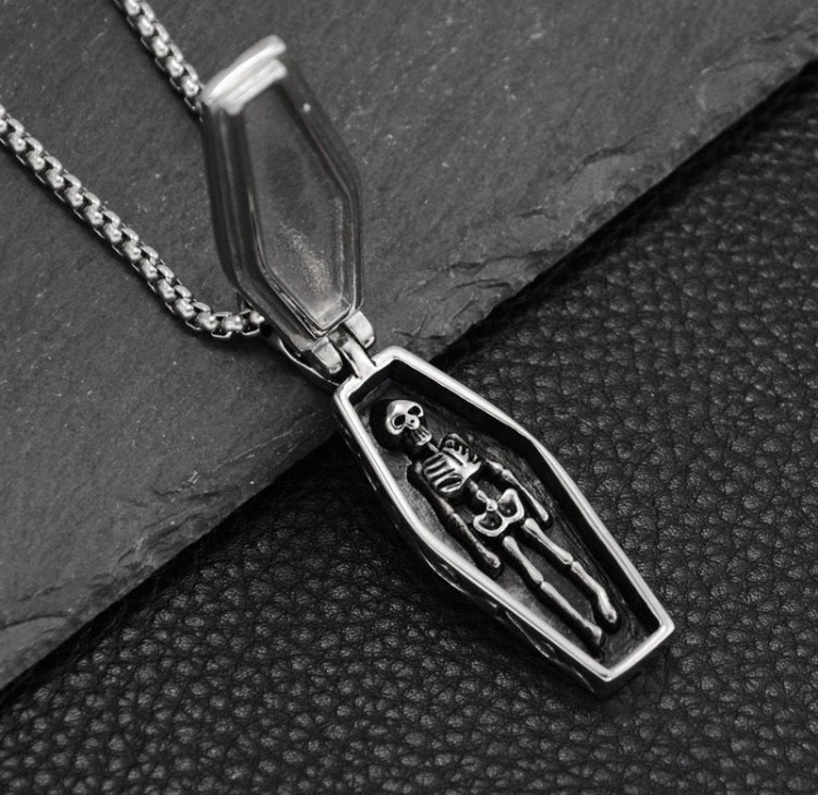 A coffin shaped pendant is open to reveal a skeleton lying embedded inside it. All are silver in colour and there is a solid looking chain attached to it.