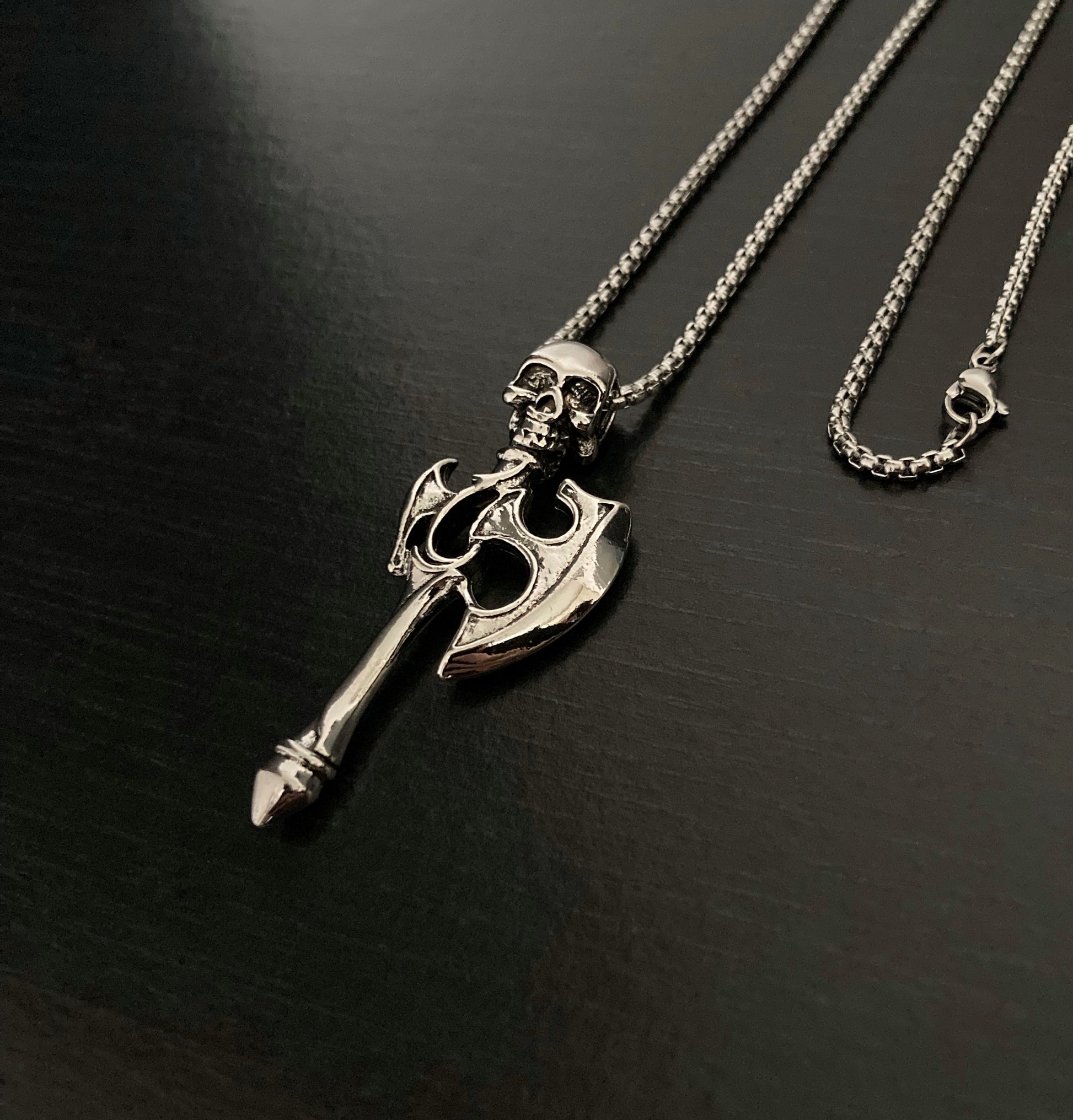 A angled view of axe pendant that features a skulls at the top of it. You can see the chain as well as the metal clasp for fastening. 