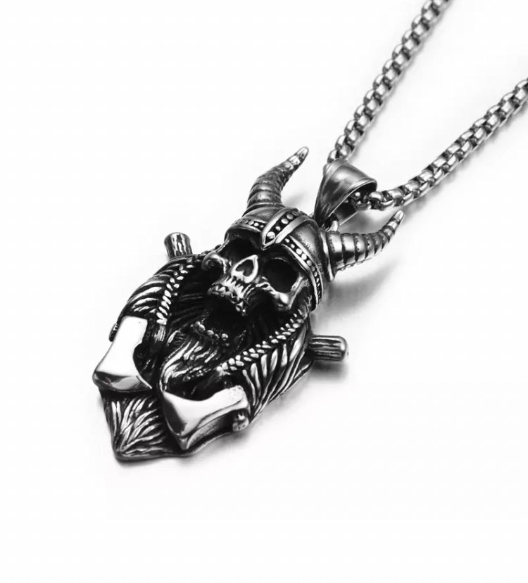 A white background hosts a skull pendant which is wearing a horned helmet and has 2 axes. Intricate detailing shows off the beauty of the skulls features with a open mouth giving it a sinister look. Silver in colour and made of stianless steel.