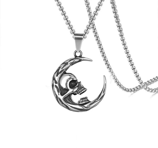 On a plain white background is a silver colour pendant that is in the shape of a crescent moon. Built into the inner curve of this is a skull face with a gaping mouth that bares teeth. Attached to this is a chain of the same colour.
