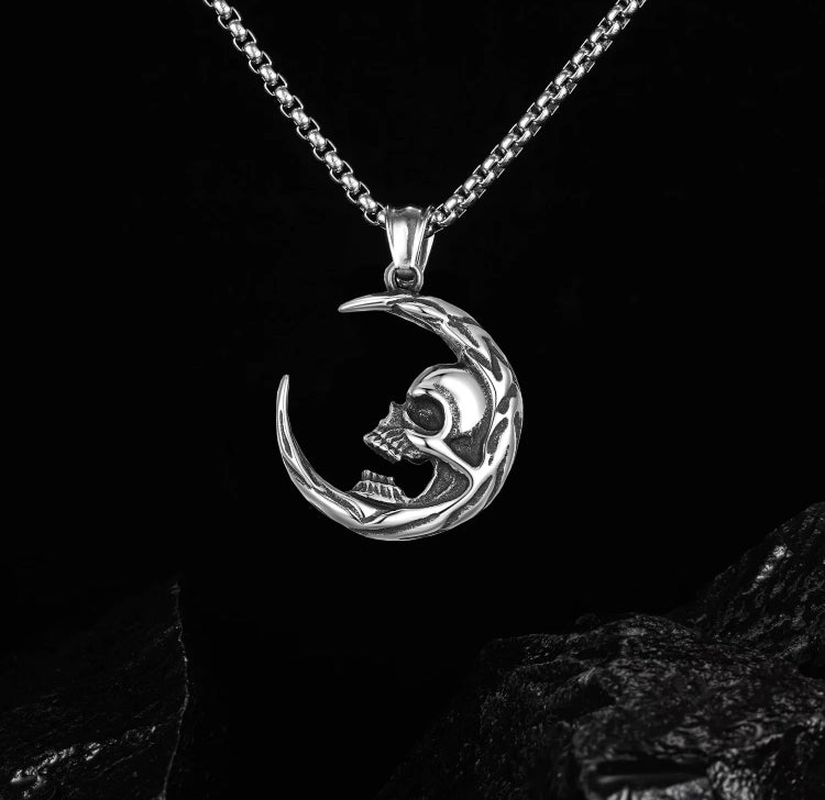 Over some rocks dangles a crescent moon necklace. This is attached to a chain and both are silver in colour. A skull face sits in the curve of the moon and looks sinister with bared teeth and hollowed out eyes.