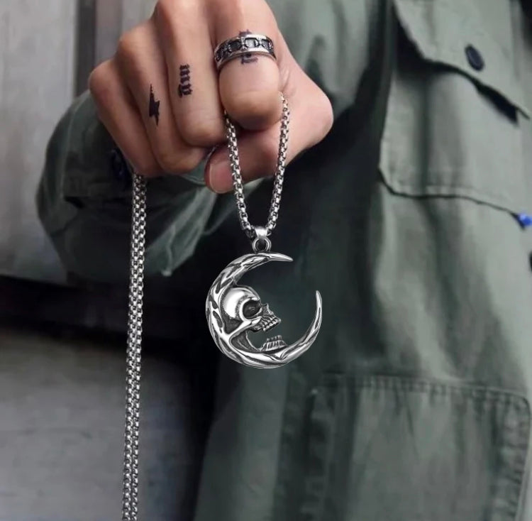 A clenched fist grips the chain of a crescent moon pendant that has a skull attached to it in the inner curve of the moon. The chain dangles down and looks to be sturdy. There is an engraved pattern on the edge of the crescent.