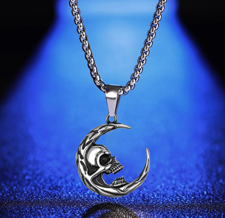 A close up of a skull in a crescent moon necklace. It is silver in colour including the chain which looks to be sturdy. There are markings on the edge of the moon and the skull has a gaping wide mouth.