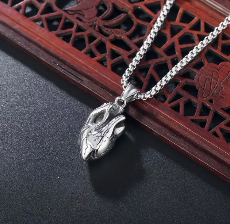 Draped over a brown patterned surface is a solid pendant in the shape of a human heart. This is a very detailed item with hollowed out holes as the valves and engravings of what look like veins on it. This is silver in colour as is the chain. 