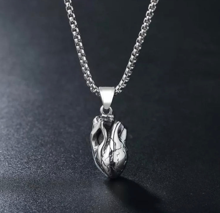 An anatomical heart pendant is dangling over a grey surface, the sturdy chain on show that is attached to it. This is a solid weighty item to hold and you can feel the holes which represent the hearts valves. Silver in colour and beautifully detailed