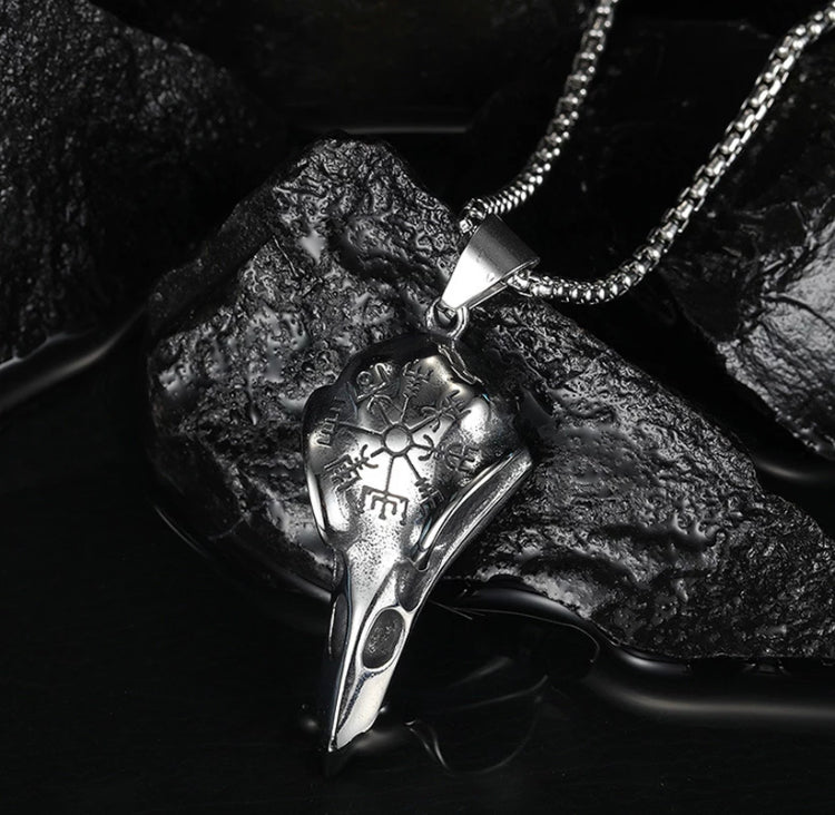 A close up view of a ravens skull pendant. A very detailed helm of awe design is etched onto the top part of the skull. Attached to this is a sturdy looking chain and the whole item is leaning against a black rock.