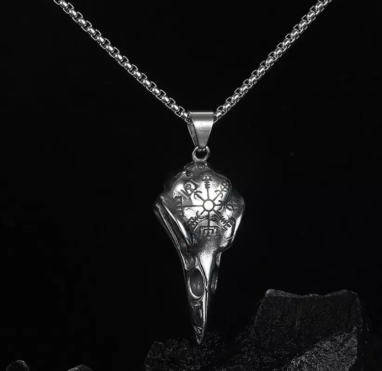 A necklace hangs over a black rock. It is silver in colour with a link chain and a pendant in the shape of a birds skull. There are hollowed out bits for the nostrils on the beak and a design engraved onto the skull itself.