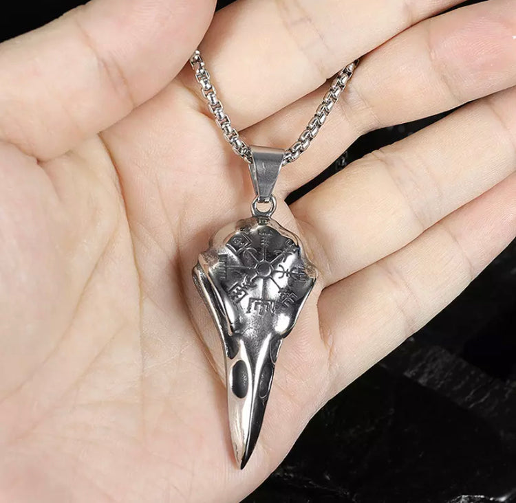 In the open palm of a hand sits a ravens skull pendant. It is lying at an angle and you can see the intricate details of the engraving and the hollowed out nostrils and beak markings.