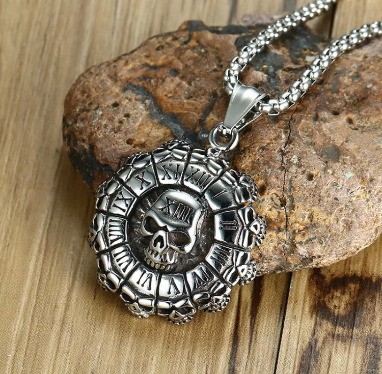 Lying across a stone is a silver coloured necklace with a round pendant attached to it. Shaped like a metal bottle cap there are     Roman numerals made to look like a clock face round the edge with a skull in the centre.