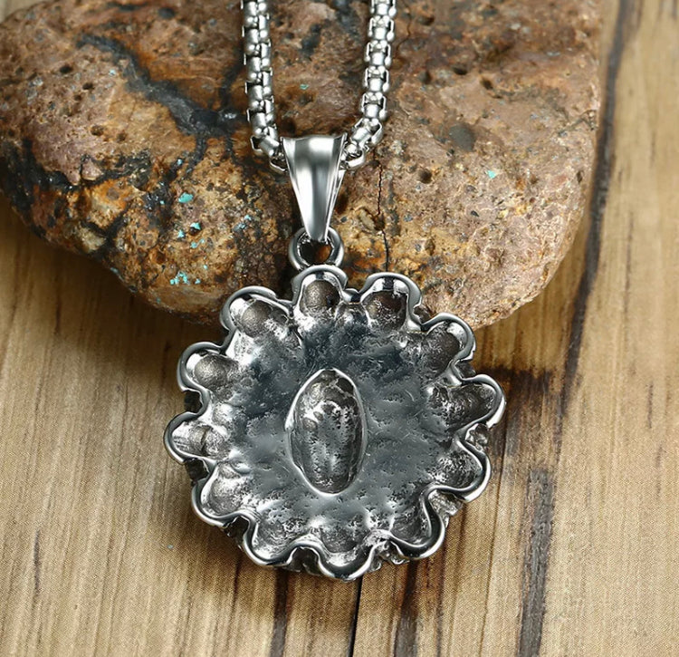 Against a rock is the reverse side of a silver coloured circular pendant. This view shows it is hollow and plain on the back of it. You can see the sturdy chain attached to the top of the round item which looks both strong and quality.