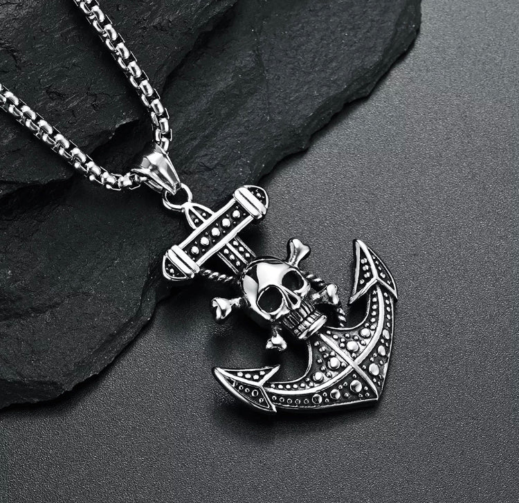 Lying on a black stone background is a stainless steel pendant with necklace. This is in the shape of an anchor with a detailed pattern on it and on the handle is a skull and crossbones design.