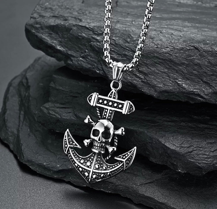 A close up view of a skull and crossbones attached to an anchor necklace. The pendant is shiny silver coloured and the anchor is decorated with circular dot patterns. The skull is situated on the top part of the anchor and is baring its teeth.