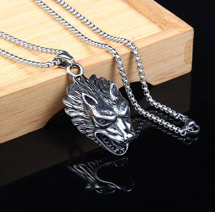 Leaning against a piece of wood is a necklace featuring a chain and a wolfs head pendant. Both are silver in colour and you can clearly see the metal clasp that fastens it together.