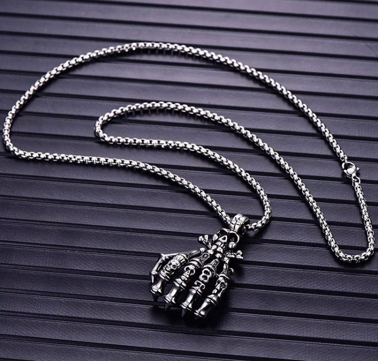 A sturdy looking chain holds a skeletons hand pendant which is detailed and features various skulls carved onto it. There is a metal fastening clasp that can be seen and it is all silver coloured.