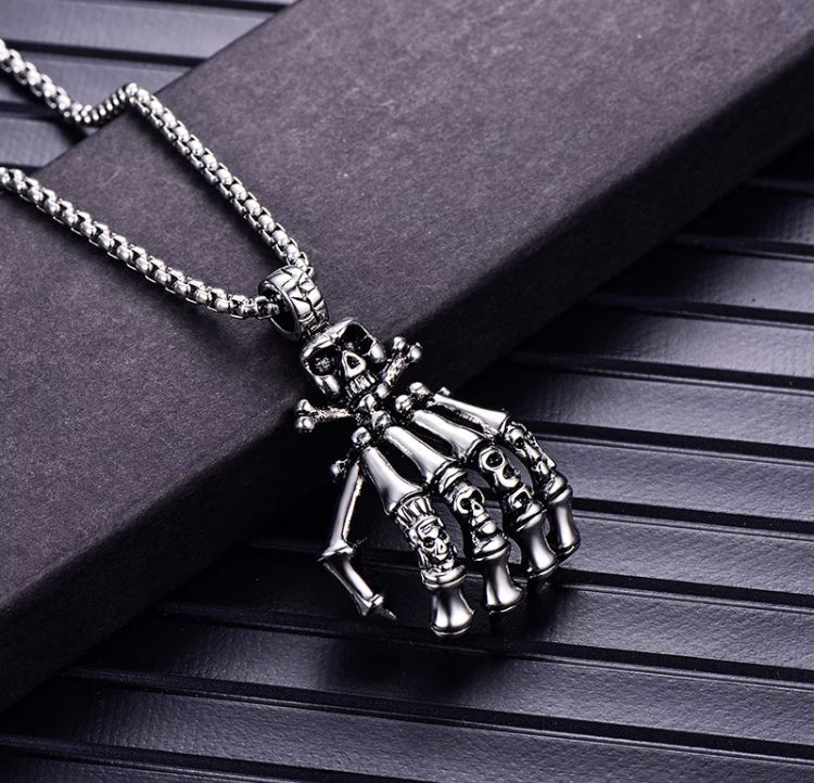 A close up view of a skeletons hand pendant. Attached to a chain with a ring the hand is very detailed with skull and crossbones at the top and the knuckles of the fingers have different carvings of skulls on each one.