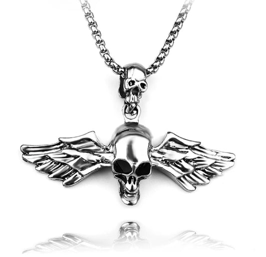 A silver coloured necklace is hanging on a white background. The chain goes through a small skull and then attached to that is a much larger skull with wings coming out of either side of it. This is very detailed and the wings feature carved patterning.