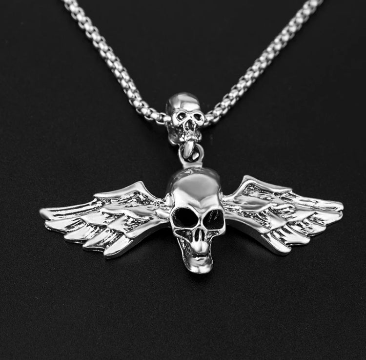 A chunky looking pendant is on a black surface. Attached to a chain via a smaller skull that sits above it this is silver in colour and shiny in appearance.