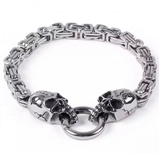On a white background sits a silver coloured bracelet. It has a link style chain and either end of that sits a skull head with a ring through the mouths holding the bracelet together.