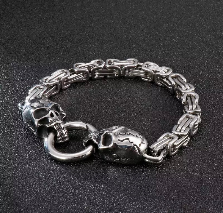 On a grey background there is a bracelet with two skulls either ends of a link chain, these are connected by a ring in the mouths. The chain links are rectangular and the whole thing is a silver colour.