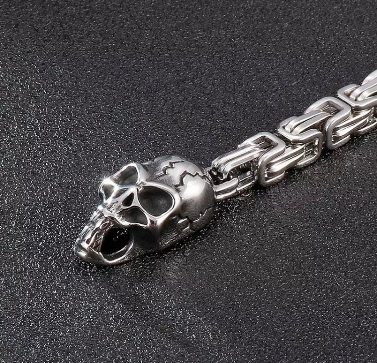 An angled view of a skull head attached to a link chain. Silver in colour the skull has detailed markings that look like cracks in it as well as facial features. It looks solid and shiny in appearance.