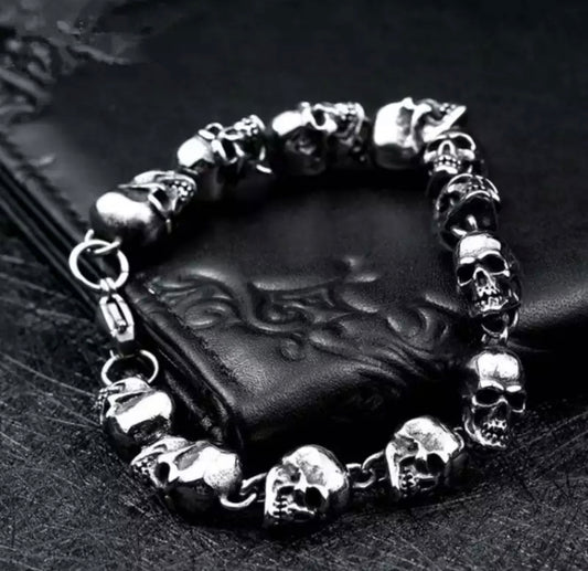 A silver coloured bracelet sits on a black wallet. It is silver in colour and features skulls heads all around it with 2 small links between each one. The skulls feature hollowed out eyes and the teeth are on show.