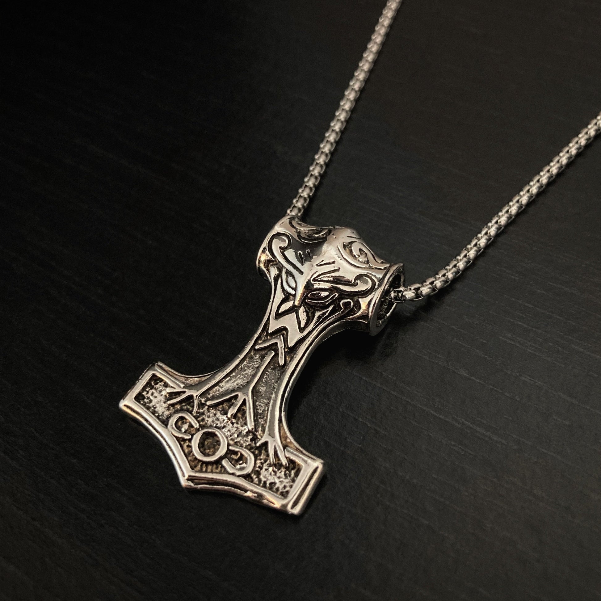 A silver coloured necklace sits on a black background which has a pendant in the shape of a viking style hammer. There is a Algiz rune on the main piece of the hammer and other markings around it. This looks solid and of good quality whilst not being too chunky.