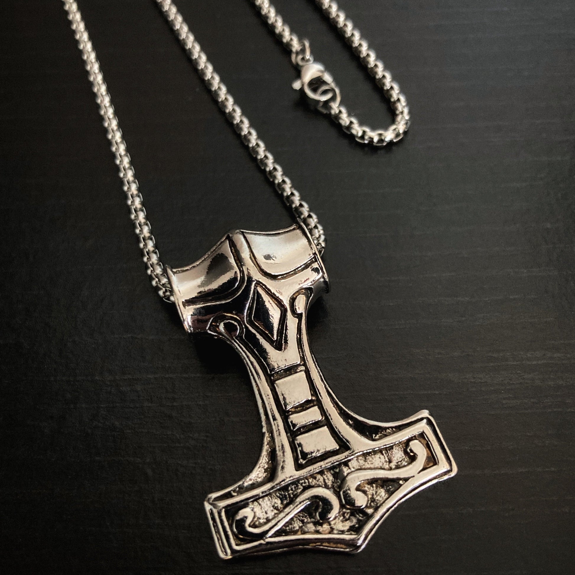 The reverse side of a pendant in the shape of Thors hammer mjolnir. There are markings engraved onto this side of the item which are intricate in detail. You can see the quality looking chain with its metal clasp.