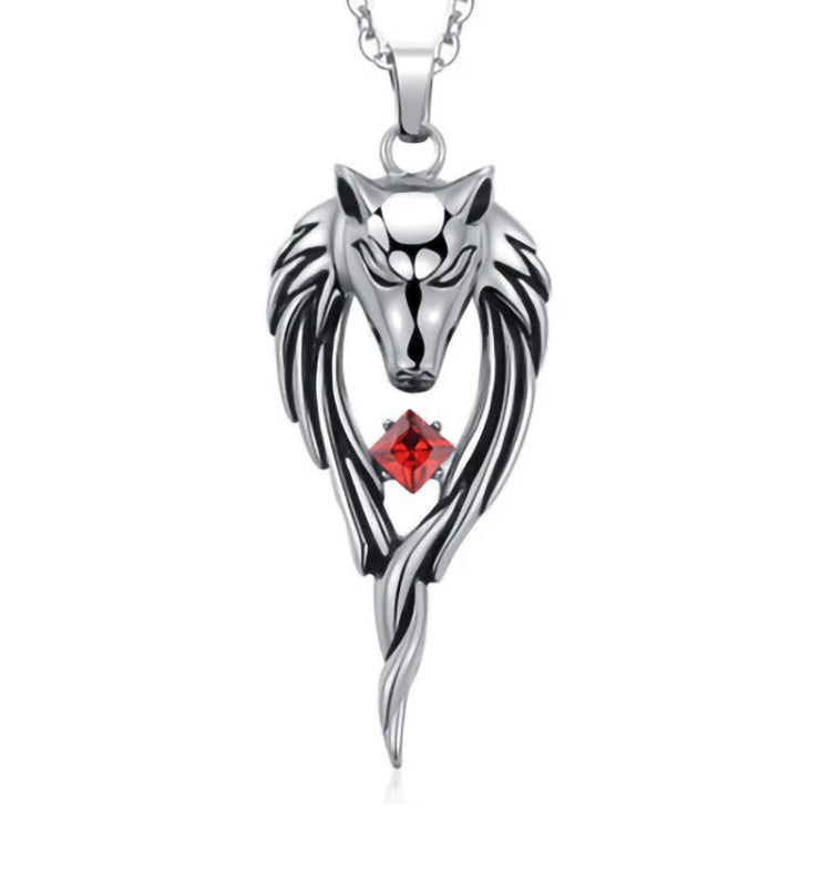 A silver coloured wolfs head pendant is hanging on a white background. Either side of the head there is detailed etched fur which hangs down and meets at the bottom. In the middle of this is a cut out piece where a red glass stone sits. 