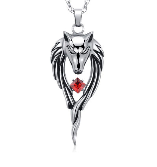 A silver coloured wolfs head pendant is hanging on a white background. Either side of the head there is detailed etched fur which hangs down and meets at the bottom. In the middle of this is a cut out piece where a red glass stone sits. 