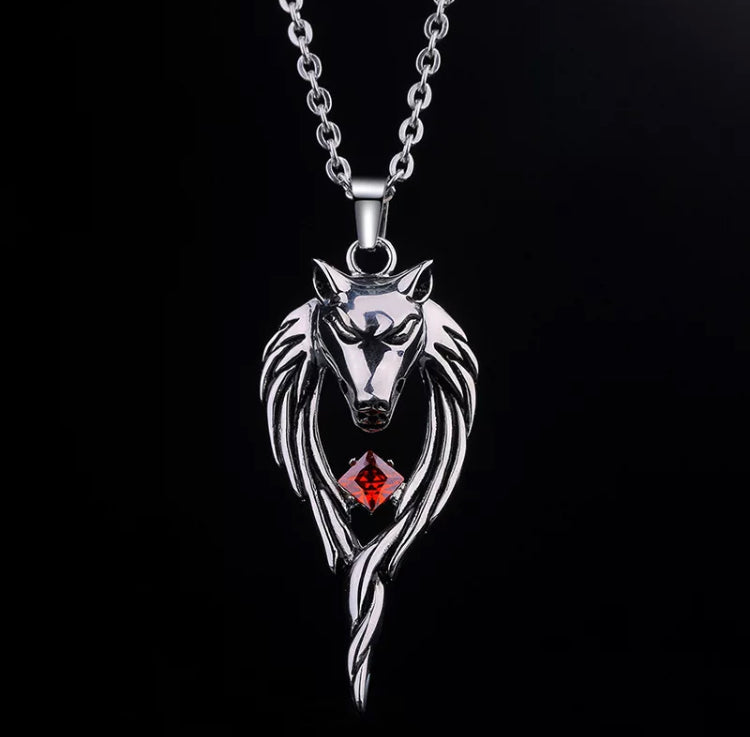 On a black background hangs a wolfs head necklace. The head is detailed in design with facial features and a long mane of fur that hangs down. Between this sits a red stone. The necklace is stainless steel and is chunky but not heavy.