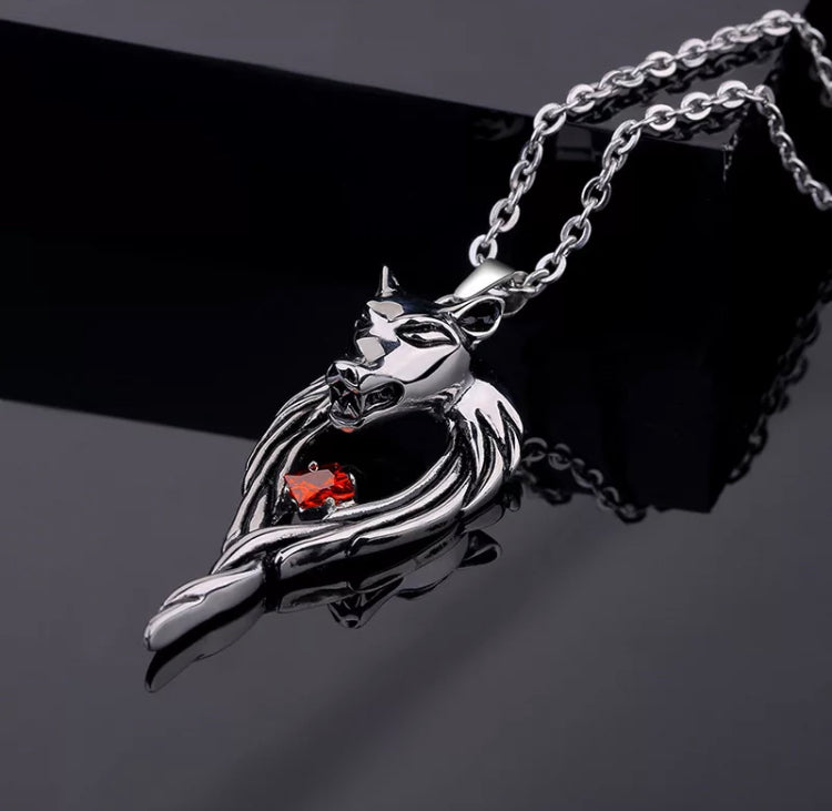 On a chain sits a wolfs head pendant. Both are silver in colour and shiny in appearance. The pendant also features a red glass stone set into the middle of the design.